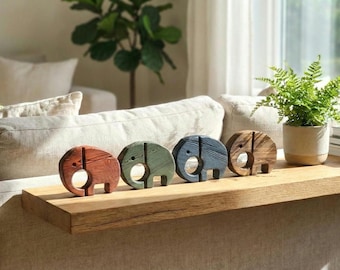 May include: Four wooden elephant figurines in shades of red, green, blue, and brown are displayed on a wooden shelf. Each elephant has a circular body with a trunk and a cutout for the eye. A fern in a beige pot sits nearby.