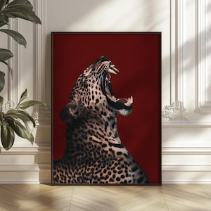 Könnte beinhalten: Gerahmter Kunstdruck mit einer Nahaufnahme eines brüllenden Leoparden vor tiefrotem Hintergrund. Das gefleckte Fell des Leoparden ist sichtbar, und sein Maul ist weit geöffnet, wobei seine Zähne zu sehen sind. Das Kunstwerk wird in einem schwarzen Rahmen präsentiert.