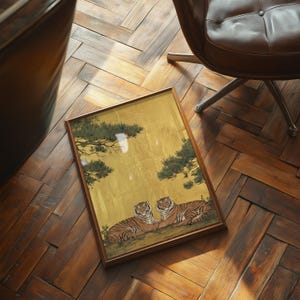 May include: A framed artwork featuring two tigers resting on a hillside, set against a gold background with green foliage. The artwork is in a wooden frame and rests on a wooden floor.