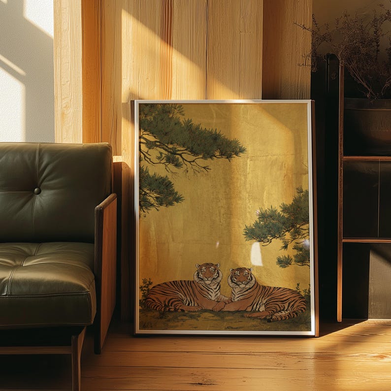 May include: Framed artwork featuring two tigers resting side-by-side. The tigers have orange and black stripes, set against a gold background with green foliage. The artwork is in a silver frame and is displayed on a wooden floor.