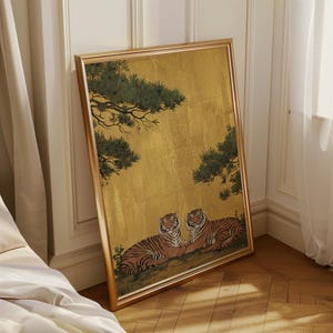 May include: Framed artwork featuring two tigers resting beneath a golden background with green tree branches. The tigers have orange and black stripes. The frame is gold-colored and leans against a wall.