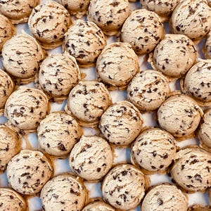 May include: A close-up of a tray of cookie dough balls, each with chocolate chips. The dough balls are arranged in rows and are a light brown color.
