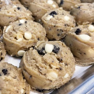May include: Close-up of a tray of cookie dough balls with white chocolate chips and Oreo cookie pieces. The dough is light brown and the chips are white.