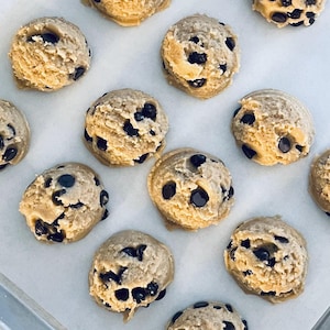 May include: Close-up of a baking sheet with 10 round cookie dough balls. The dough is a light brown color and has chocolate chips throughout. The dough balls are arranged in a random pattern on the baking sheet.
