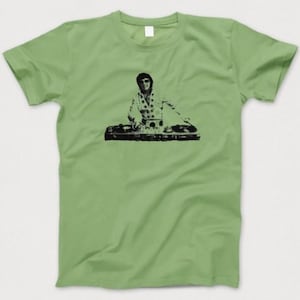 May include: A pale green t-shirt with a black graphic of a DJ at work. The DJ is wearing sunglasses and a patterned shirt. The t-shirt has a crew neck and short sleeves. A casual garment.