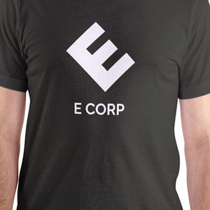 Puede incluir: Camiseta gris oscuro con un logotipo "E" blanco estilizado y el texto "E CORP" debajo. La camiseta es de cuello redondo y parece estar hecha de un material suave.
