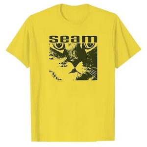 Könnte beinhalten: Gelbes T-Shirt mit einer schwarz-weißen Grafik eines Katzenkopfes und dem Wort "seam" darüber. Das Design ist zentriert, wobei die Augen der Katze ein Schlüsselelement sind. Die Schriftart ist fett und blockartig.
