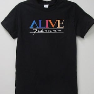 Op de afbeelding: Zwart T-shirt met de tekst "ALIVE films". Het woord "ALIVE" is in blauw, oranje en geel, met "films" in een handgeschreven lettertype eronder. Een eenvoudig, stijlvol ontwerp.