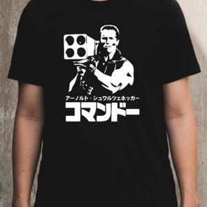 May include: Black t-shirt with a white graphic of a man holding a weapon. The graphic includes Japanese text and the name "Arnold Schwarzenegger". The shirt has a crew neck.