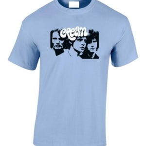 May include: Light blue t-shirt featuring a black and white graphic of the band Cream. The word "cream" is above the band members' faces. The shirt has short sleeves and a crew neck.