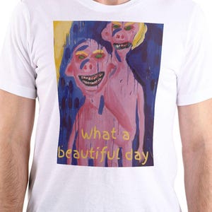 Può includere: T-shirt bianca con un dipinto astratto e colorato di due figure con tratti esagerati e occhi verdi. L'opera d'arte include il testo "What a beautiful day" in giallo.