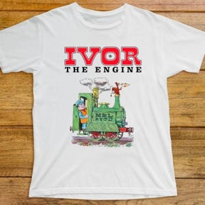 Puede incluir: Camiseta blanca con el texto "IVOR THE ENGINE" en rojo. Debajo, una colorida ilustración de un tren verde con una persona en el asiento del conductor. El tren emite humo.