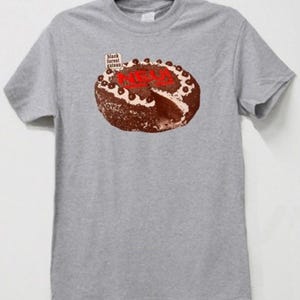 Può includere: T-shirt grigia con una grafica di una torta Foresta Nera, con la parola "NEU" in rosso. La maglietta ha anche il testo "black forest gateau" in un piccolo riquadro. Alla torta manca una fetta.