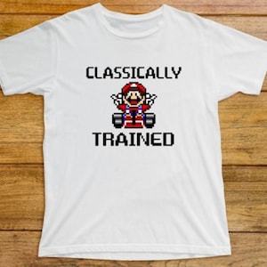 May include: White t-shirt featuring the text "CLASSICALLY TRAINED" and a pixelated image of Mario in a kart. The image is in red, blue, and black. The shirt is on a wooden surface.