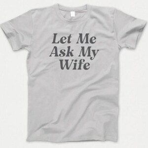 Könnte beinhalten: Ein hellgraues T-Shirt mit dem Text "Let Me Ask My Wife" in Dunkelgrau. Das Shirt hat kurze Ärmel und einen Rundhalsausschnitt. Der Text ist mittig auf der Vorderseite des Shirts platziert.