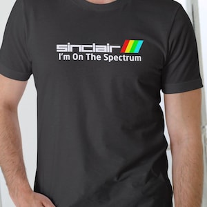 Puede incluir: Una camiseta negra con texto blanco que dice "Sinclair I'm On The Spectrum" y una franja de colores arcoíris.