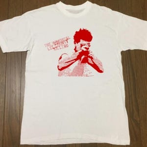 Op de afbeelding: Wit t-shirt met een rode afbeelding van een persoon en de tekst "THE IMMORTAL LEE SCRATCH PERRY KILLERS". De afbeelding is een gestileerde illustratie. Het shirt is gemaakt van een zacht materiaal.