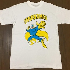 May include: White t-shirt featuring a cartoon superhero with the text "BANANAMAN" in yellow. The superhero is depicted in blue and yellow, with a muscular build and a dynamic pose, suggesting action and strength. The shirt is made of a soft, comfortable material.