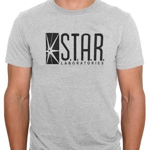 May include: A light gray t-shirt featuring the text "STAR LABORATORIES" in black. The design includes a geometric star graphic. The shirt is short-sleeved and made of a soft material.