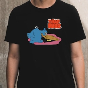 Puede incluir: Camiseta negra con un personaje de dibujos animados azul, sosteniendo una tabla de madera sobre una trampilla rosa y marrón. El texto "TRAP DOOR" es rojo y naranja. Una prenda informal.