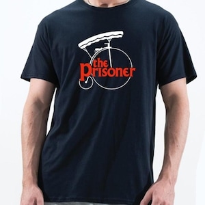 Könnte beinhalten: Marineblaues T-Shirt mit einem weißen und roten Grafikdesign eines Hochrads und dem Text "the Prisoner". Das Design befindet sich auf der Vorderseite des Shirts.