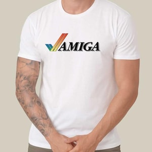 May include: White t-shirt featuring the word "AMIGA" in black, with a colourful logo above it. The logo is a stylized checkmark with red, orange, yellow, green, and blue stripes. The shirt has short sleeves.