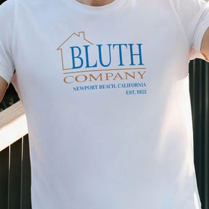 以下が含まれることがあります： 白のTシャツ。青と茶色で「BLUTH COMPANY」の文字と家のグラフィックがプリントされています。その下に「NEWPORT BEACH, CALIFORNIA EST. 1953」と書かれています。クルーネックのスタイルです。