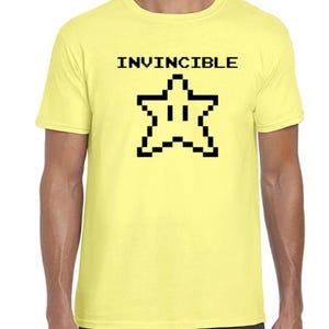 May include: A pale yellow t-shirt featuring the word "INVINCIBLE" in black, block letters above a black, pixelated star design. The shirt has a crew neck and short sleeves.