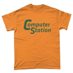 Puede incluir: Camiseta naranja con el texto "Computer Station" en verde. La camiseta es de cuello redondo y parece estar hecha de algodón o un tejido similar. El texto está centrado en la parte delantera de la camiseta.