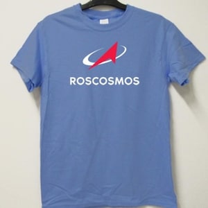 Puede incluir: Una camiseta azul claro con el logotipo de ROSCOSMOS. El logotipo es una flecha roja dentro de un diseño curvo blanco, con la palabra "ROSCOSMOS" en letras blancas debajo. La camiseta está colgada en una percha negra.