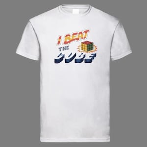 May include: White t-shirt with the text "I BEAT THE CUBE" in red and blue, featuring a graphic of a Rubik's Cube. The design has a retro, vintage style. The shirt is made of a soft, comfortable material.