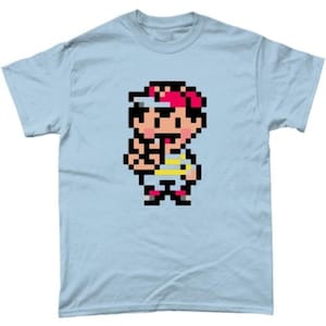 Peut inclure: T-shirt bleu clair avec un graphisme pixélisé d'un personnage aux cheveux rouges et une casquette blanche et rouge. Le personnage porte une chemise rayée jaune et blanche et un short foncé. Le t-shirt est en matière souple.