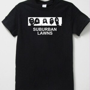 May include: Black t-shirt featuring a white rectangular graphic with four stylized faces and the text "SUBURBAN LAWNS" in white block letters. The shirt has short sleeves and a crew neck.
