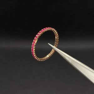 May include: A delicate gold band ring with a row of small, bright red gemstones. The ring is held by tweezers against a black background.