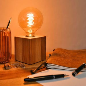 Könnte beinhalten: Ein warmer Schreibtischaufbau mit einer Holzlampe und einer großen Edison-Glühbirne, einem gerippten orangefarbenen Glas-Stifthalter mit Bleistiften, einem ledergebundenen Notizbuch, Stiften und Papier. Gemütliche Arbeitsatmosphäre.