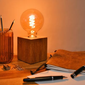 Könnte beinhalten: Ein warmer Schreibtisch mit einer Holzlampe und einer leuchtenden Glühbirne. Ein geriffelter, bernsteinfarbener Glas-Stifthalter enthält Bleistifte. Ein ledergebundenes Notizbuch, Stifte und ein Spitzer sind ebenfalls vorhanden.