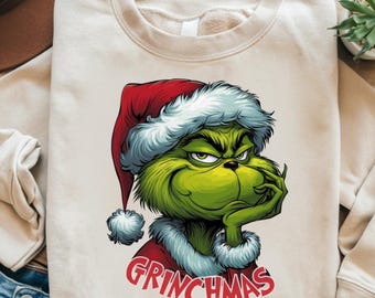 May include: Cream-colored sweatshirt featuring a cartoon Grinch character wearing a Santa hat and red coat. The Grinch is green with a grumpy expression, and the word "GRINCHMAS" is printed in red below.