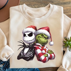 Könnte beinhalten: Cremefarbenes Sweatshirt mit einer Grafik von Jack Skellington und dem Grinch, beide mit Weihnachtsmützen. Jack trägt einen schwarz-weißen Anzug, während der Grinch ein rot-weiß gestreiftes Outfit trägt. Die Charaktere posieren mit verschränkten Armen.