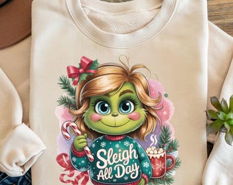 May include: Cream-colored sweatshirt featuring a cartoon character with green skin, blonde hair, and blue eyes. The character is wearing a teal sweater with the text "Sleigh All Day" and holding a candy cane. A red mug with marshmallows is also visible.