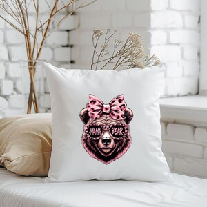 Pu&ograve; includere: Cuscino bianco con un orso rosa e marrone che indossa occhiali da sole che dicono "Mama Bear". L'orso ha un fiocco rosa sulla testa.