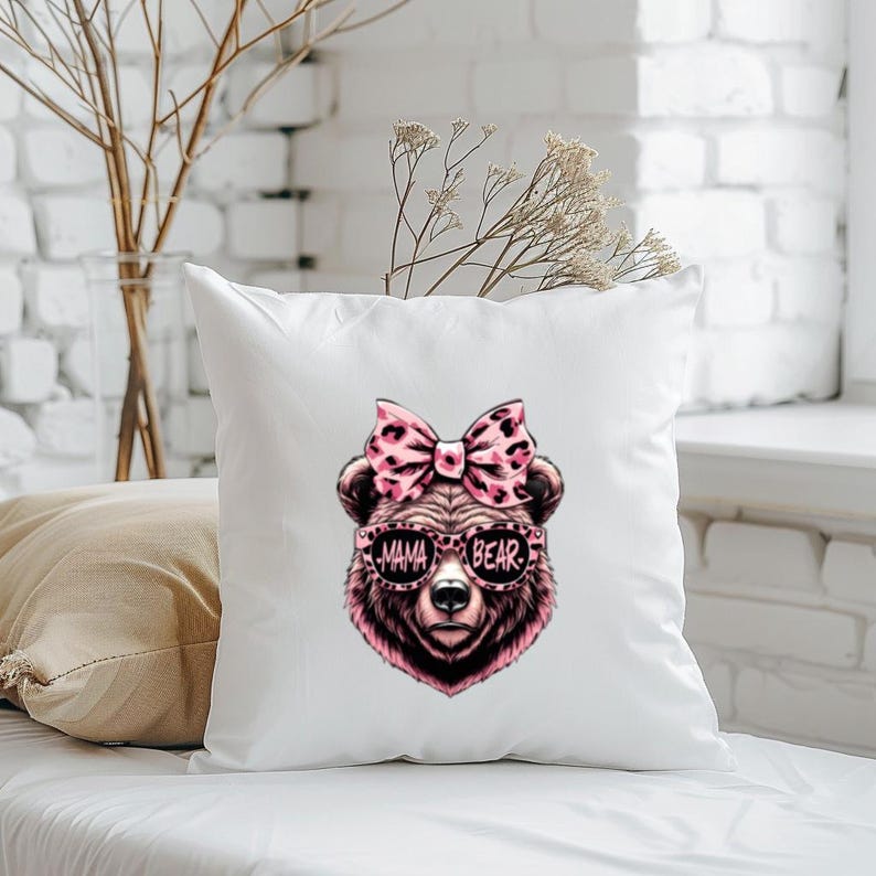 Pu&ograve; includere: Cuscino bianco con un orso rosa e marrone che indossa occhiali da sole che dicono "Mama Bear". L'orso ha un fiocco rosa sulla testa.