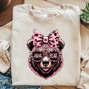 Pu&ograve; includere: Una felpa beige chiaro con un orso rosa che indossa occhiali da sole che dicono "Mama Bear" e un fiocco rosa con stampa leopardata.