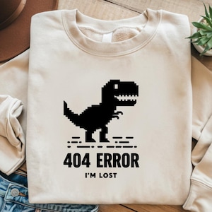 May include: A beige sweatshirt with a black pixelated dinosaur graphic and the text "404 ERROR I'M LOST".