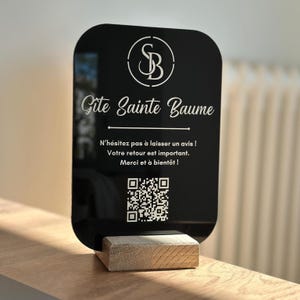 Può includere: Un cartello in acrilico nero con angoli arrotondati, con il testo "Gite Sainte Baume" e un logo. Il cartello è montato su una base di legno. Il testo aggiuntivo include un invito all'azione e un codice QR.