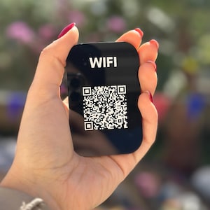 Può includere: Una scheda rettangolare nera con la parola "WIFI" in testo bianco sopra un codice QR. La scheda è tenuta in una mano con smalto rosso, con uno sfondo sfocato di verde e rosa.
