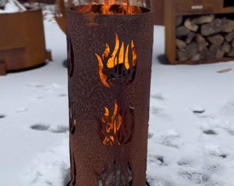 May include: A rusty metal fire pit with a cutout design of flames. The fire pit is burning with orange flames and is sitting on a snowy surface.