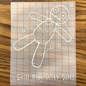 May include: A transparent sticker featuring a white outline of a voodoo doll with pins. The sticker has an orange grid pattern and the text "I Still Play With Dolls" in white.