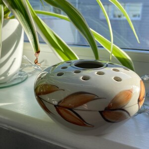 May include: A white ceramic flower frog with multiple holes for arranging stems. The oval-shaped vase features brown leaf designs and is displayed on a windowsill with greenery.