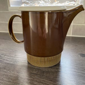 May include: A brown ceramic teapot with a cream lid and a handle. The teapot has a spout and a decorative base with vertical lines. The teapot is on a wooden surface.