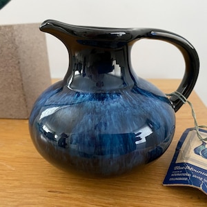 May include: A ceramic jug with a glossy finish. The jug has a dark blue and black colour scheme, with a rounded body and a handle. A tag is attached to the handle. The jug is likely used for serving beverages.